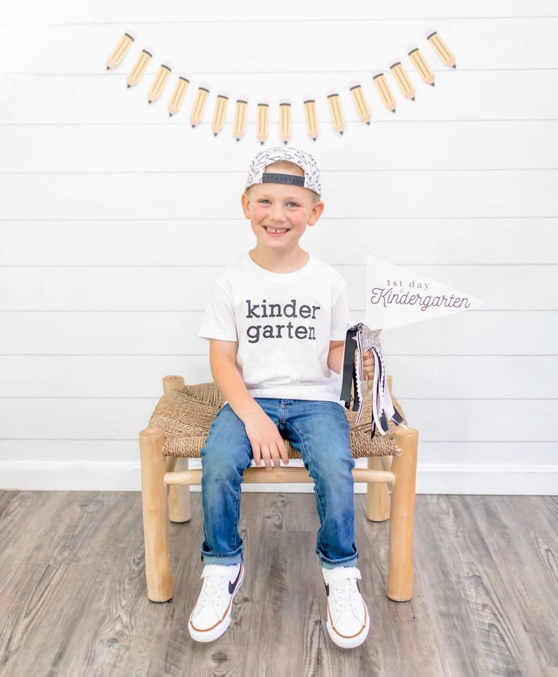 May include: A young boy wearing a white t-shirt with the text "kinder garten" printed on it, blue jeans, and a black and white patterned baseball cap. He is holding a white sign that says "1st day Kindergarten" and is sitting on a wooden stool.