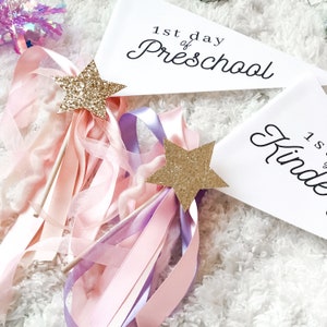 May include: Two white pennants with black text. The top pennant says "1st day of Preschool". The bottom pennant says "1st day of Kindergarten". Two gold glitter star wands with pink and purple ribbons are in the foreground.
