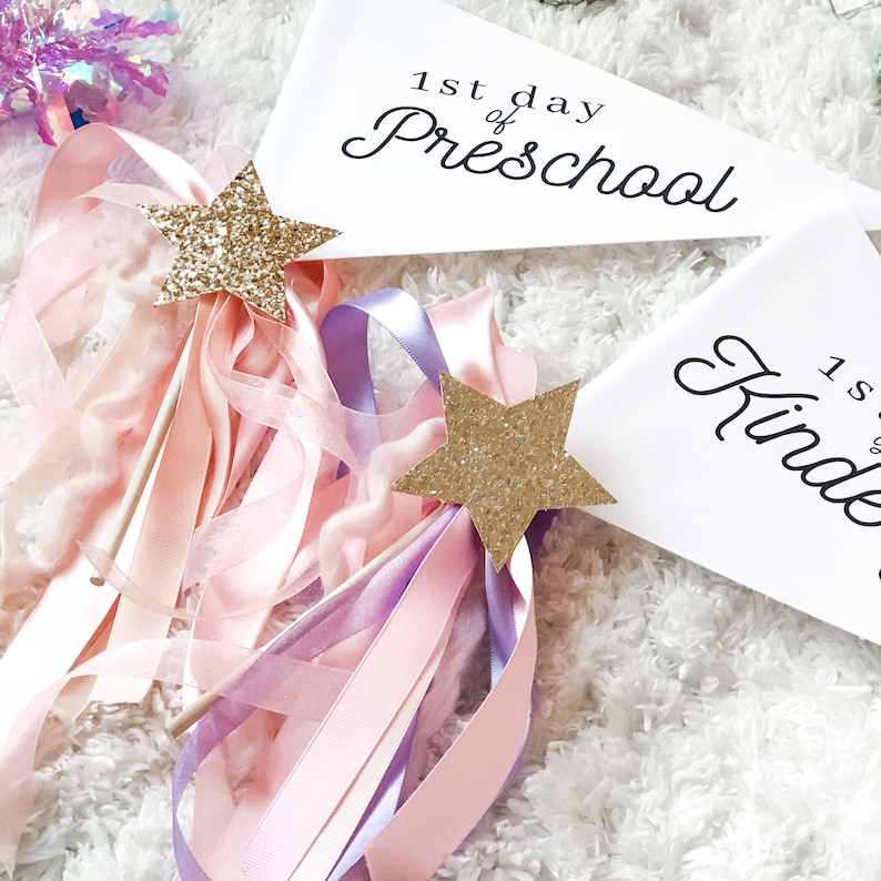 May include: Two white pennants with black text. The top pennant says "1st day of Preschool". The bottom pennant says "1st day of Kindergarten". Two gold glitter star wands with pink and purple ribbons are in the foreground.