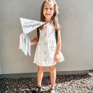 May include: A young girl in a white dress holds a sign that says "first day of kindergarten" with a sparkly star and colourful streamers attached. She is smiling and wearing black shoes.