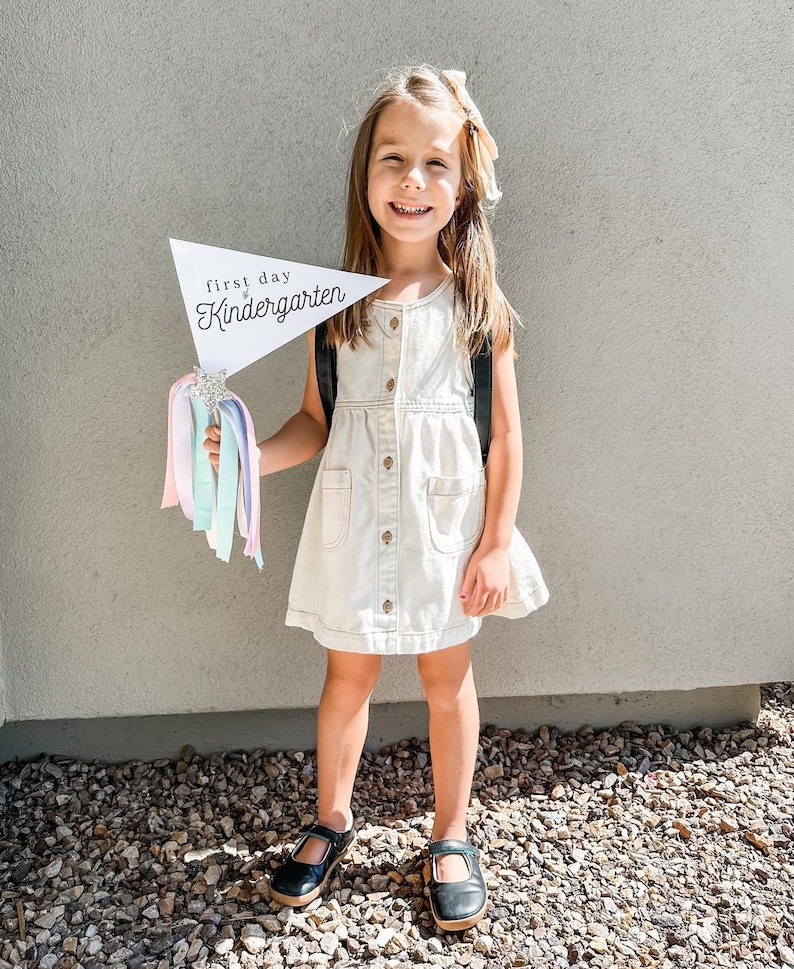May include: A young girl in a white dress holds a sign that says "first day of kindergarten" with a sparkly star and colourful streamers attached. She is smiling and wearing black shoes.