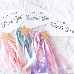May include: Three white pennants with black text, each reading "1st day of First Grade", "1st day of Second Grade", and "1st day of Fourth Grade". The pennants are decorated with gold glitter stars and pink, purple, and blue ribbons.