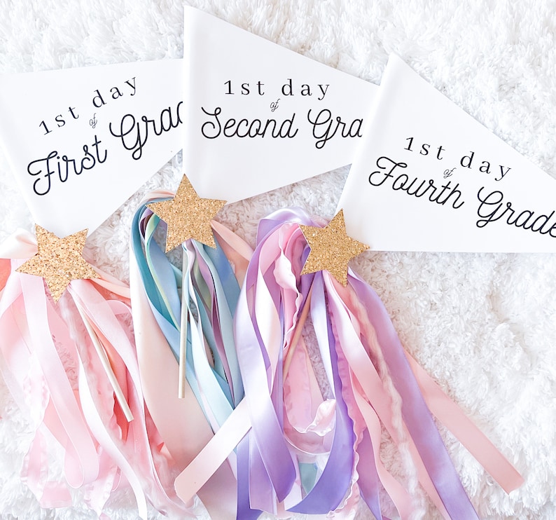 May include: Three white pennants with black text, each reading "1st day of First Grade", "1st day of Second Grade", and "1st day of Fourth Grade". The pennants are decorated with gold glitter stars and pink, purple, and blue ribbons.