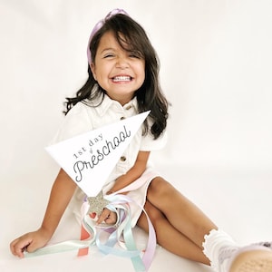 May include: A young girl wearing a white dress and a purple headband holds a white sign that says "1st day of Preschool" with a gold glitter star. She is smiling and holding a wand with colourful ribbons.