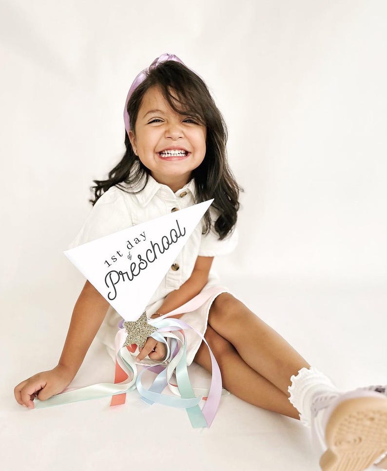 May include: A young girl wearing a white dress and a purple headband holds a white sign that says "1st day of Preschool" with a gold glitter star. She is smiling and holding a wand with colourful ribbons.