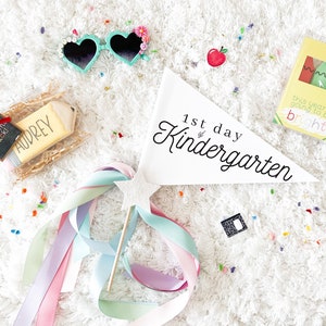 May include: A white pennant with the text "1st day of Kindergarten" in black script. There are several other items in the image, including a pencil-shaped cookie, a welcome back sign, a pair of heart-shaped sunglasses, a box of crayons, and a wand with colourful ribbons.
