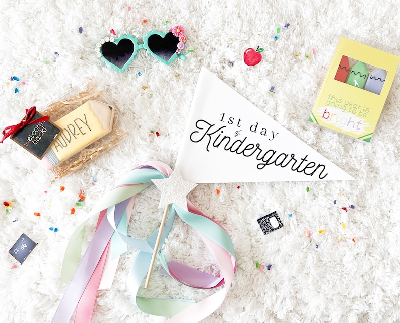 May include: A white pennant with the text "1st day of Kindergarten" in black script. There are several other items in the image, including a pencil-shaped cookie, a welcome back sign, a pair of heart-shaped sunglasses, a box of crayons, and a wand with colourful ribbons.