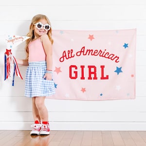 May include: A young girl wearing white sunglasses and a pink tank top stands in front of a pink banner with blue and white stars. The banner reads "All American Girl" in red letters. She is wearing a blue and white gingham skirt and red sneakers.