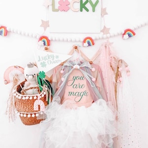 May include: A pink and white tutu with a pink shirt that says "you are magic" hanging on a wooden hanger. The background is white with a white beaded garland with rainbow and star decorations. There is a sign that says "Lucky Charm" with a green shamrock.