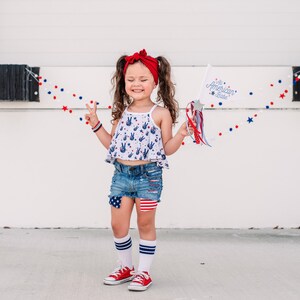 May include: A young girl wearing a red headband, a white tank top with blue peace signs, denim shorts with red, white, and blue patches, white socks with blue stripes, and red sneakers. She is holding a white flag with the text "The American Babe" and a red, white, and blue streamer. She is standing in front of a white wall with a red, white, and blue garland.