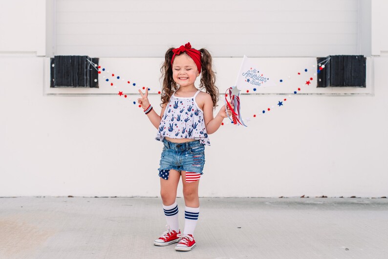 May include: A young girl wearing a red headband, a white tank top with blue peace signs, denim shorts with red, white, and blue patches, white socks with blue stripes, and red sneakers. She is holding a white flag with the text "The American Babe" and a red, white, and blue streamer. She is standing in front of a white wall with a red, white, and blue garland.