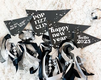 May include: Black and white New Year's Eve party decorations with the text "Pop Fizz Clink", "Happy New Year", and "Hello 2025" on black pennant flags. Silver glitter star wands with black and white ribbons.