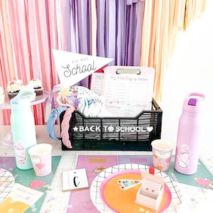 May include: A back to school party table setting with pink, purple, and yellow streamers. The table is decorated with a black and white grid tablecloth, confetti, and a black basket with the words "Back to School" on it. There are two water bottles, one blue and one pink, with names on them. There are also two cups with hearts on them. There is a plate with a rainbow design and a small box with a flag that says "1st Grade" on it.
