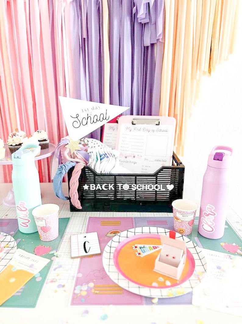 May include: A back to school party table setting with pink, purple, and yellow streamers. The table is decorated with a black and white grid tablecloth, confetti, and a black basket with the words "Back to School" on it. There are two water bottles, one blue and one pink, with names on them. There are also two cups with hearts on them. There is a plate with a rainbow design and a small box with a flag that says "1st Grade" on it.