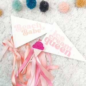 May include: Two white pennant flags with pink text. One flag says "Beach Babe" and the other says "Ice Cream Queen". A pink and white glitter ice cream cone is in the center of the image.