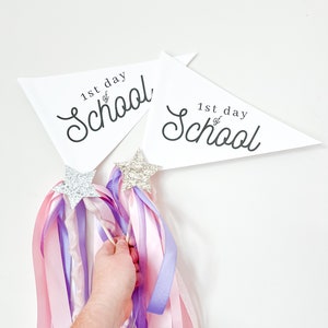 May include: Two white triangular pennants with the text "1st day of School" in black cursive writing. Two sparkly silver star wands with pink and purple ribbons.