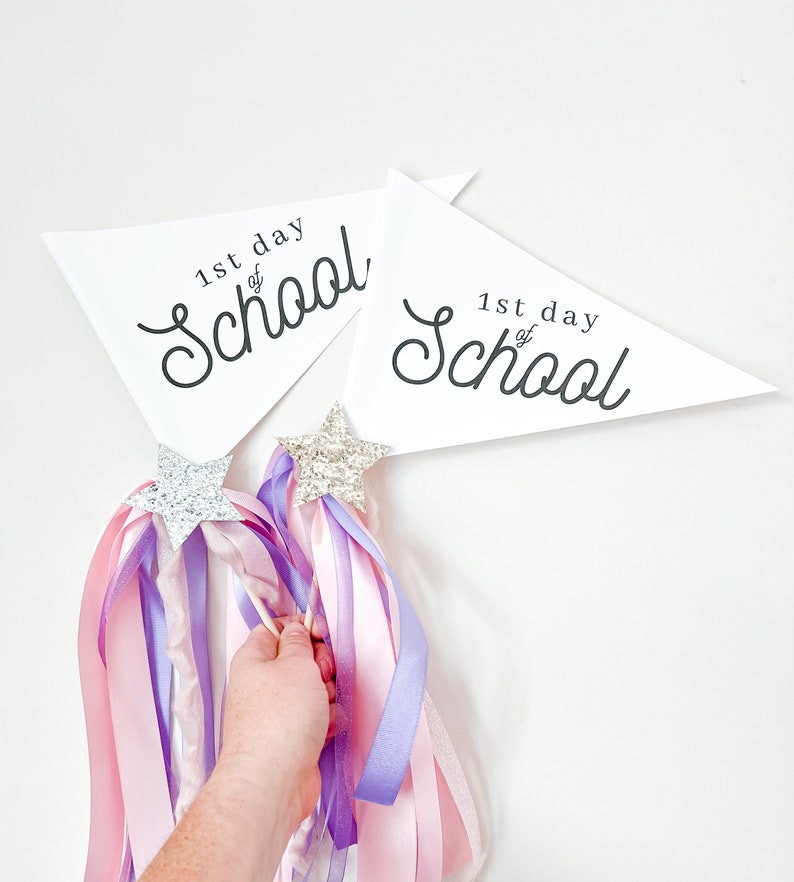 May include: Two white triangular pennants with the text "1st day of School" in black cursive writing. Two sparkly silver star wands with pink and purple ribbons.