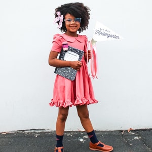 May include: A young girl wearing a pink dress, glasses, and a pink bow in her hair. She is holding a notebook and a sign that says "1st day of Kindergarten".