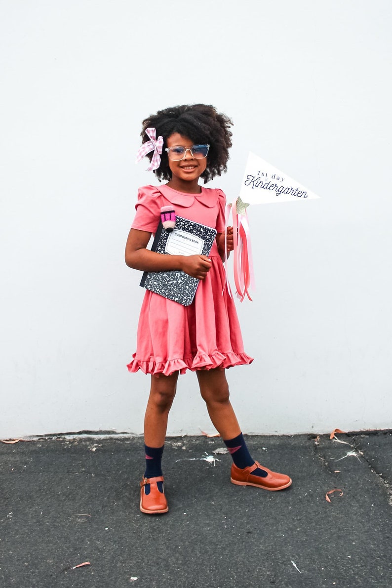 May include: A young girl wearing a pink dress, glasses, and a pink bow in her hair. She is holding a notebook and a sign that says "1st day of Kindergarten".