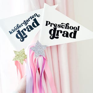 Puede incluir: Dos banderines triangulares blancos con texto negro, uno dice "kindergarten grad" y el otro dice "preschool grad". Los banderines están unidos a serpentinas rosas y moradas con estrellas doradas y plateadas de brillantina.