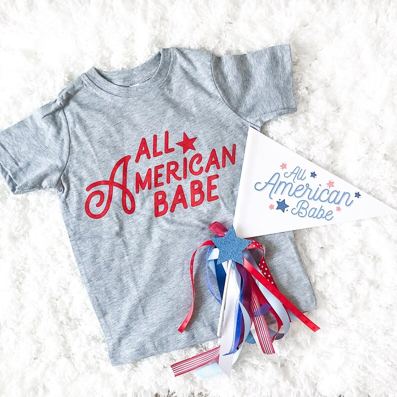 May include: A gray t-shirt with the text "All American Babe" in red lettering and a red star. A white pennant with the same text in blue lettering and stars is also shown. A red, white, and blue streamer is included.
