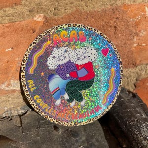 Peut inclure: Un autocollant rond avec un fond holographique représentant deux personnages de dessins animés qui s'embrassent. L'autocollant porte le texte "ACAB" en couleurs arc-en-ciel en haut et "ALL COUPLES ARE BEAUTIFUL" sur le bord. La bordure a un motif léopard.