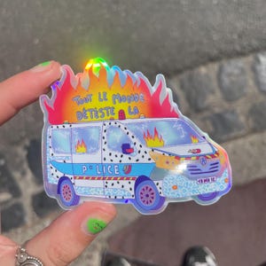Peut inclure: Un autocollant holographique représentant une camionnette blanche avec des accents bleus et un motif léopard, marquée P*LICE. Le véhicule est entouré de flammes orange et rouges, avec le texte TOUT LE MONDE DÉTESTE LA écrit en bleu au-dessus.