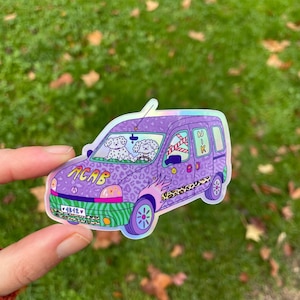 Può includere: Un adesivo olografico di un furgone viola con un motivo leopardato. Il furgone ha due cani nei sedili anteriori e la scritta "ACAB" sulla parte anteriore. L'adesivo è tenuto da una mano su uno sfondo verde.
