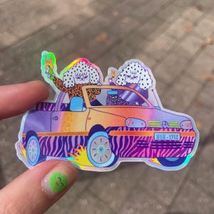 Peut inclure: Un autocollant holographique représentant deux personnages aux cheveux blancs et taches noires conduisant un cabriolet à la carrosserie dégradée violette et orange, avec des panneaux inférieurs zébrés et des flammes jaunes, plaque NIK-1512.