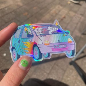 Peut inclure: Un autocollant holographique représentant une voiture bleu clair et violette avec des flammes sur le côté. Deux chiens blancs aux poils bouclés sont à l'intérieur, l'un au volant et l'autre tenant une bouteille verte. La plaque indique NIK 1312.