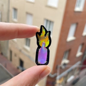 Peut inclure: Un autocollant holographique en forme de bouteille avec des flammes. L'autocollant a un contour noir et est tenu entre deux doigts. Les flammes sont jaunes et orange, et la bouteille est violette et bleue.