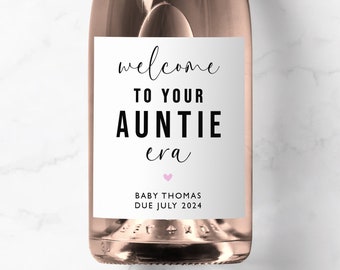 May include: A rose-colored champagne bottle with a white label. The label reads "welcome TO YOUR AUNTIE era" in a stylish font, with a small pink heart. Below, it says "BABY THOMAS DUE JULY 2024".