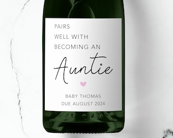 May include: A white label on a green bottle of champagne with the text "Pairs well with becoming an Auntie" and a pink heart. The label also includes the text "Baby Thomas Due August 2024".