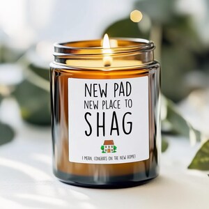 May include: A lit brown glass candle jar with a white label that reads "NEW PAD, NEW PLACE TO SHAG". The candle has a gold-colored lid and a small house graphic. The text "I MEAN, CONGRATS ON THE NEW HOME!" is below the house graphic.