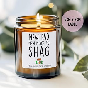 May include: A lit candle in a brown glass jar with a white label. The label reads "NEW PAD, NEW PLACE TO SHAG" with a house graphic and "I MEAN, CONGRATS ON THE NEW HOME!". A pink label indicates the label size is 5cm x 6cm.