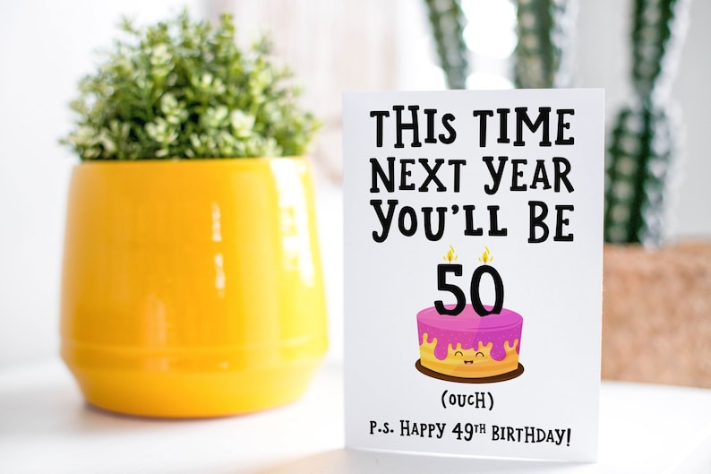 May include: A white card with black text that reads "This time next year you'll be 50 (ouch)".  The card also has a pink and yellow cake with a smiling face and the number 50 on top.  The text at the bottom of the card reads "P.S. Happy 49th Birthday!"