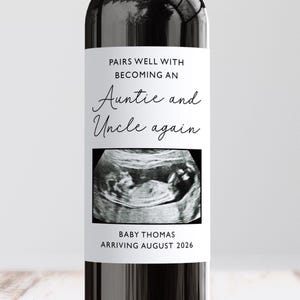 May include: A wine bottle with a custom label announcing a pregnancy. The label reads "Pairs well with becoming an Auntie and Uncle again" with an ultrasound image. The bottom of the label says "Baby Thomas arriving August 2026."