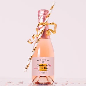 May include: A small bottle of pink sparkling beverage with a gold foil neck wrap and a gold ribbon tied around the neck. A white and gold striped paper straw is tucked into the ribbon. The label reads: BRIDE TO BE, Charlotte's Hen Do, 19TH SEPTEMBER, LONDON.