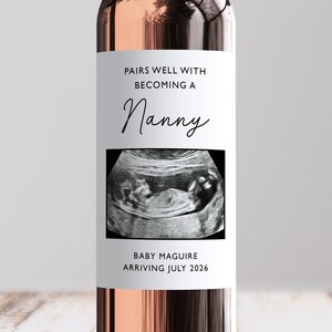 May include: A wine bottle with a white label. The label reads "PAIRS WELL WITH BECOMING A Nanny" above an ultrasound image. Below the image, it says "BABY MAGUIRE ARRIVING JULY 2026".