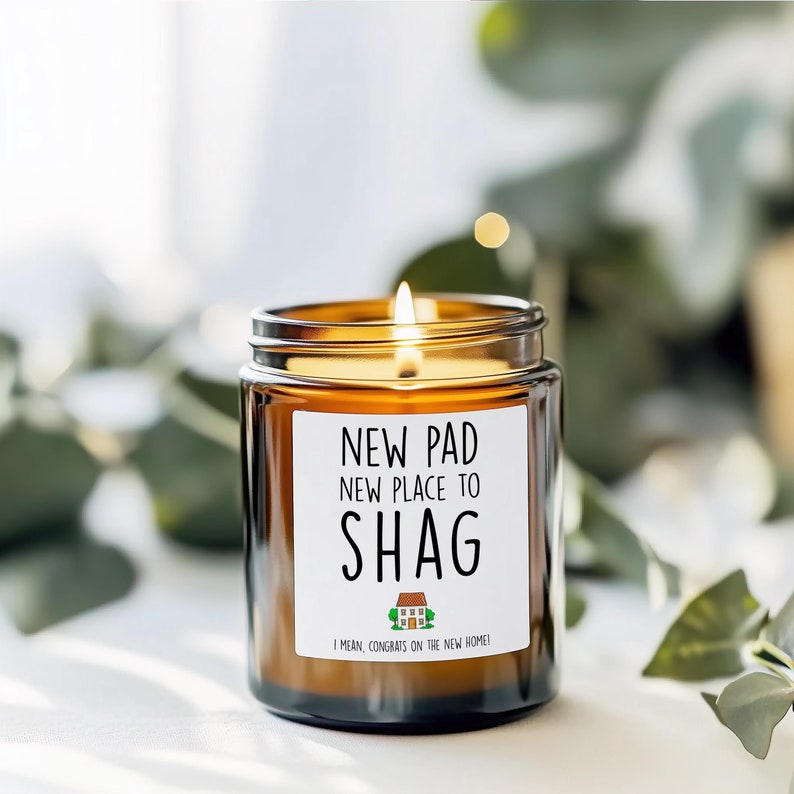 May include: A lit brown glass candle jar with a white label that reads "NEW PAD, NEW PLACE TO SHAG". The candle has a gold-colored lid and a small house graphic. The text "I MEAN, CONGRATS ON THE NEW HOME!" is below the house graphic.