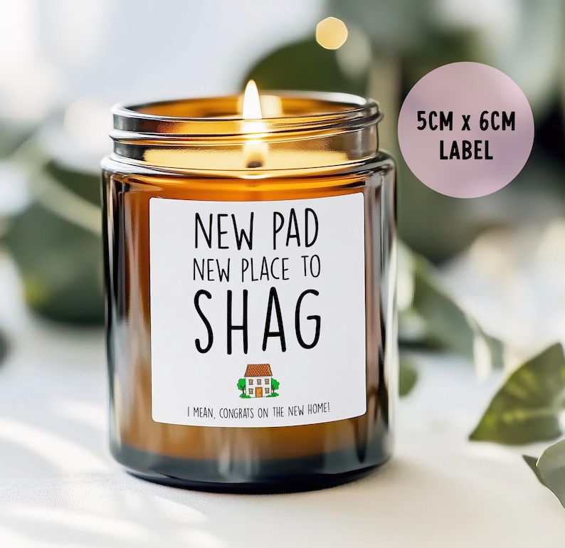 May include: A lit candle in a brown glass jar with a white label. The label reads "NEW PAD, NEW PLACE TO SHAG" with a house graphic and "I MEAN, CONGRATS ON THE NEW HOME!". A pink label indicates the label size is 5cm x 6cm.