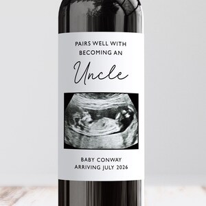May include: A wine bottle with a custom label. The label says "Pairs well with becoming an Uncle" above an ultrasound image. Below the image, it says "Baby Conway arriving July 2026."