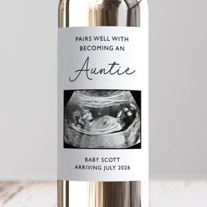 May include: A clear wine bottle with a white label. The label says "PAIRS WELL WITH BECOMING AN Auntie" in black script. Below is a grayscale sonogram image. The bottom of the label says "BABY SCOTT ARRIVING JULY 2026".