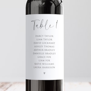 May include: A white table number card with the text "Table 1" in elegant script. Below, a list of names is printed in a simple, readable font. A small heart symbol is at the bottom. The card is set against a dark background.