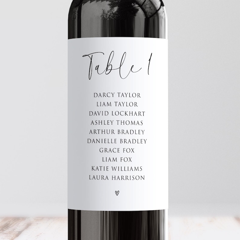 May include: A white table number card with the text "Table 1" in elegant script. Below, a list of names is printed in a simple, readable font. A small heart symbol is at the bottom. The card is set against a dark background.