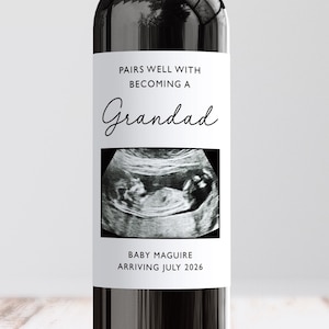May include: A wine bottle with a custom label announcing a new grandad. The label includes the text "PAIRS WELL WITH BECOMING A Grandad" and an ultrasound image. The bottom of the label reads "BABY MAGUIRE ARRIVING JULY 2026."