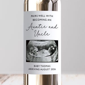 May include: A clear wine bottle with a label stating "Pairs well with becoming an Auntie and Uncle." The label includes a sonogram image and the text "Baby Thomas arriving August 2026."