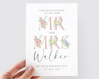 May include: A white wedding card with the text "CONGRATULATIONS TO THE NEW MR AND MRS Walker ON YOUR WEDDING DAY 21ST JULY 2025". The words "MR" and "MRS" are in pink with floral accents. The card is held by a hand.