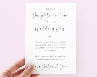 Puede incluir: Una tarjeta rectangular blanca con texto impreso en negro que dice: TO OUR Daughter in Law ON YOUR Wedding Day. Debajo hay un pequeño icono de corazón, un mensaje de bienvenida a la familia y la firma final: Love Julia & Jon.