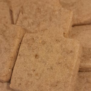 May include: A close-up shot of several square-shaped, light brown cookies. The cookies have a slightly rough texture and appear to be freshly baked. They are arranged on a white plate, suggesting a snack or dessert.